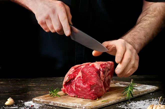 mains découpant un morceau de viande rouge sur une planche garnie de romarin